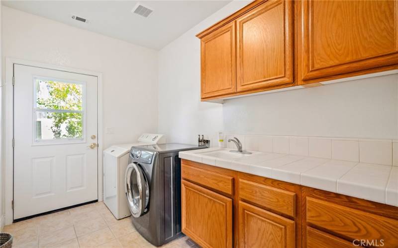 Spacious laundry room features tile countertops, deep sink, considerable cabinets and a door leading out to a patio.