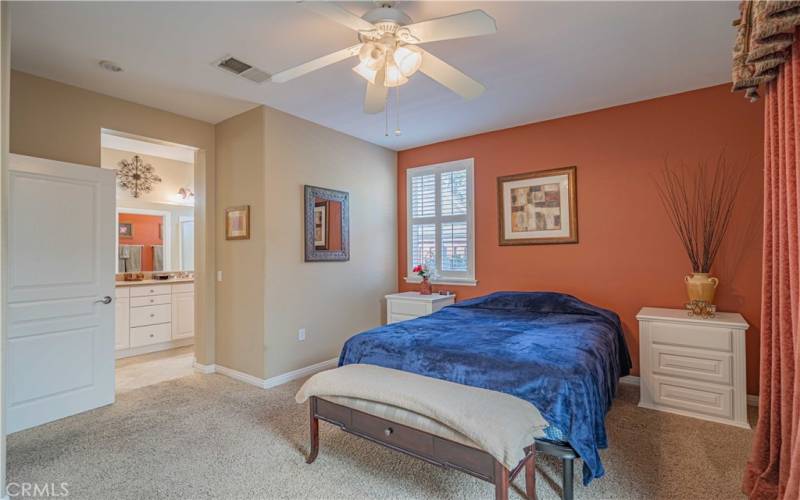 Another primary bedroom has carpet, ceiling fan, bed and white nightstands that match everything. All furniture is staying! Window with white plantation shutters. Sliding glass door with custm drapes on the right also stays.