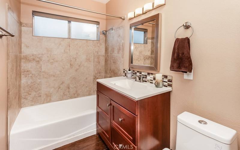 Guest bathroom with wood-grained vanity with integrated basin, framed dressing mirror, designer lighting, custom tiled tub/shower, dual flush commode and privacy window.