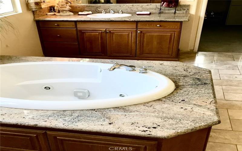 Primary bathroom has separate sinks on each side of a spa tub. Granite countertops and travertine floors