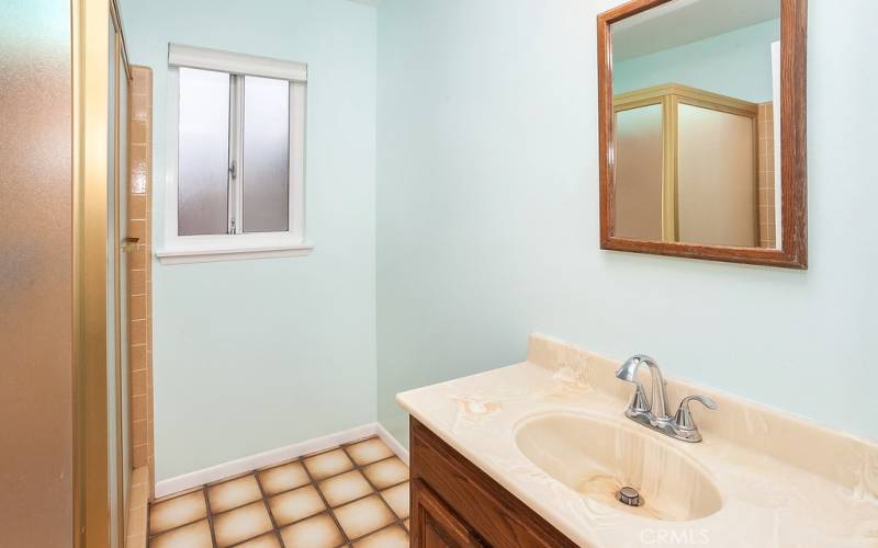 Second bathroom is freshly painted with a wood-grained vanity, framed dressing mirror, new light fixture, glass enclosed tiled step-in shower, privacy window, and tiled floors.