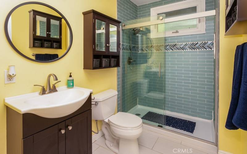 Remodeled guest bathroom with wood-grained vanity, dressing mirror, designer lights, custom subway tiled glass-enclosed step in shower with decorative inlay, dual shower heads, privacy window and custom tiled floors.