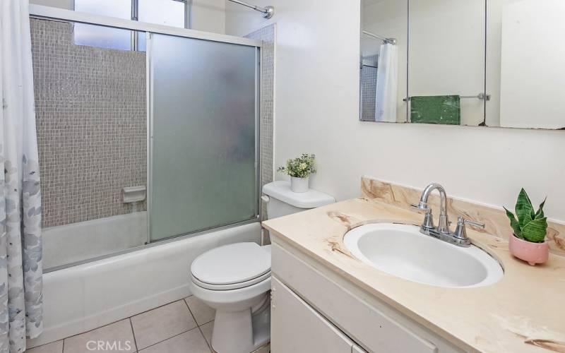 Full guest bathroom with vanity, dressing mirror, tiled tub/shower, privacy window and tiled floors.