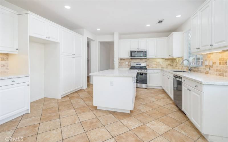 The kitchen features a quartz countertops, a center, white cabinetry, tumbled stone backsplash, and stainless steel appliances.