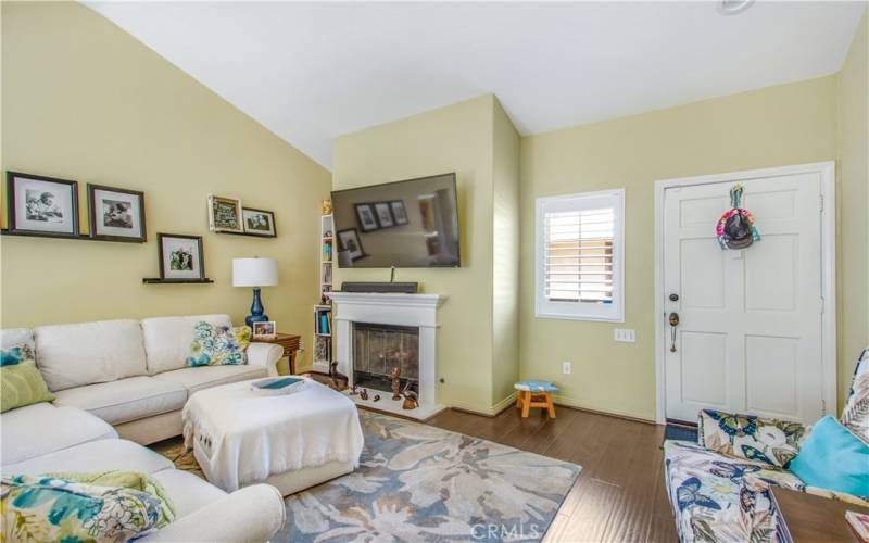 Living room including vaulted ceiling, gas fireplace and mantle, shutter window and main entry.