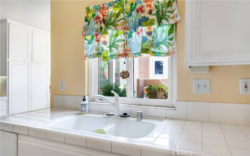 Bright white tile kitchen countertop, dual sink and dual pane sliding window to allow for light to flood into kitchen.