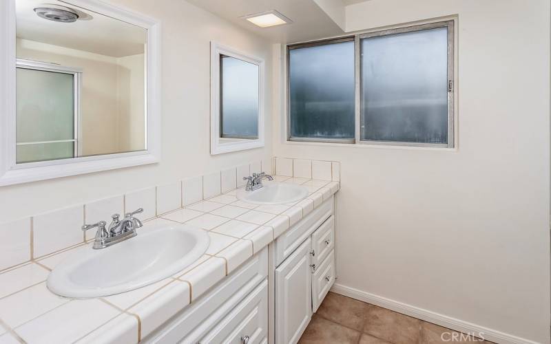 The freshly painted upstairs guest bathroom offers a white vanity, tile countertops, dual basins, large dressing mirror, glass-enclosed tub/shower, privacy window and tile floors.