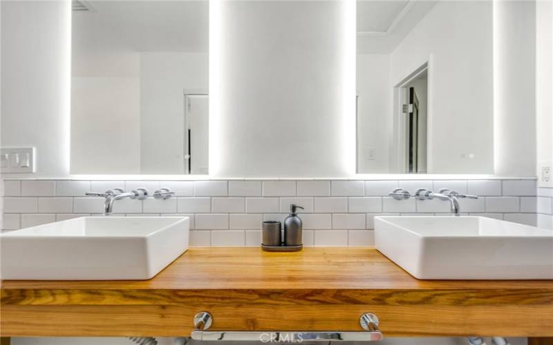 Frontal view of ensuite bathroom wooden countertop, dual sinks, back lit vanity mirrors and subway tile.