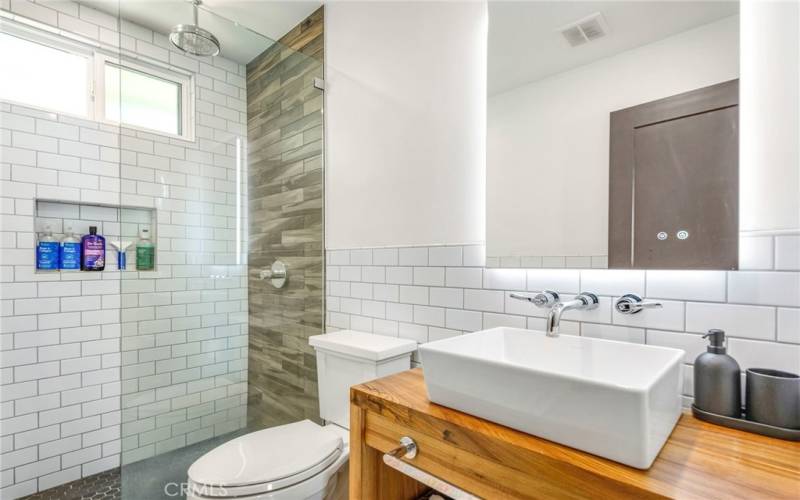 Guest bathroom, including rain shower, subway tiles, wooden counter space, back lit vanity mirror and porcelain sink.