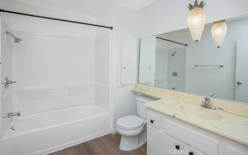 Full guest bathroom with white vanity with integrated basin, classic drop-down lighting, wall-to-wall dressing mirror, and tub/shower and tiled floors.