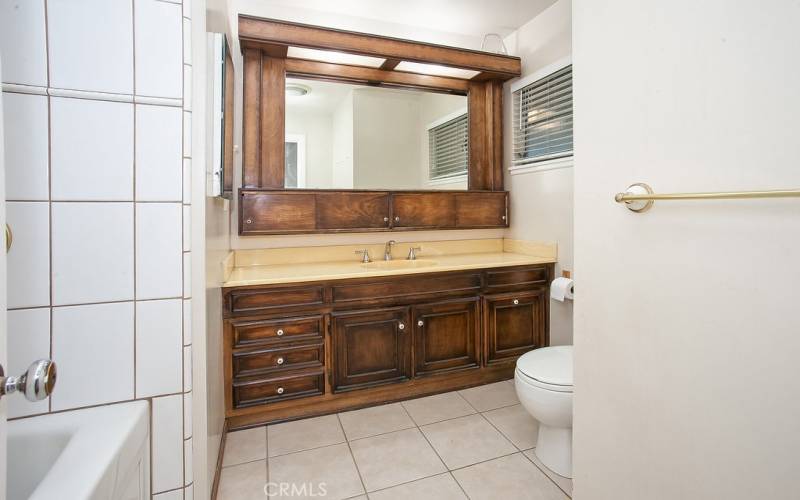 Full guest bathroom with wood-grained vanity, large dressing mirror with storage, tiled jetted tub/shower, privacy window and tile floors.