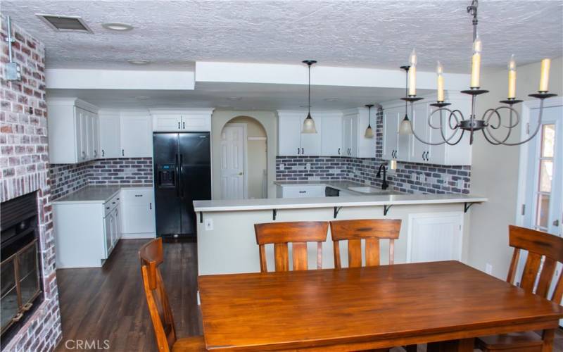 Dining area or family room with fireplace in the foreground.  Background: Kitchen with Quartz counters, modern white cabinets, custom backsplash, breakfast bar and stainless/black appliances.