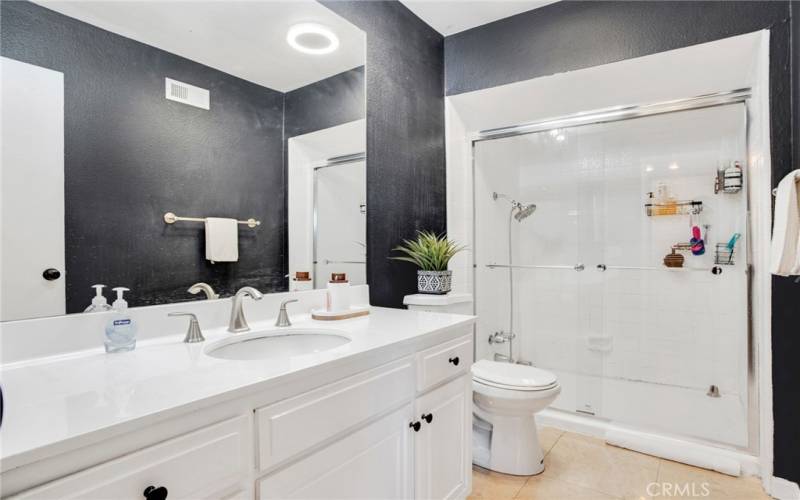 Private en suite primary bathroom with white vanity, undermount basin, dressing mirror, and modern lighting.  Separate room with additional white vanity, granite countertops, undermount basin, dressing mirror, glass enclosed tub/shower, commode and tile floors.