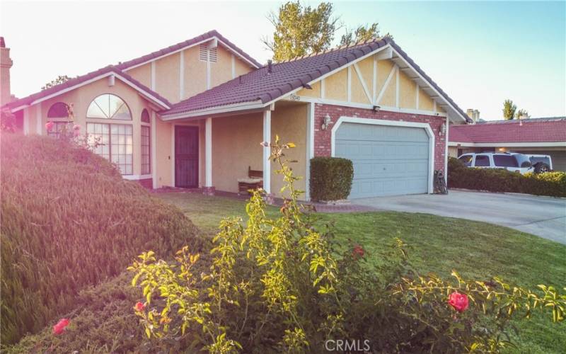 The front of the home showing grass and mature landscaping as well as brick walk way under the covered entry.  There is a tile roof and brick facade and the front door and garage are painted to match.