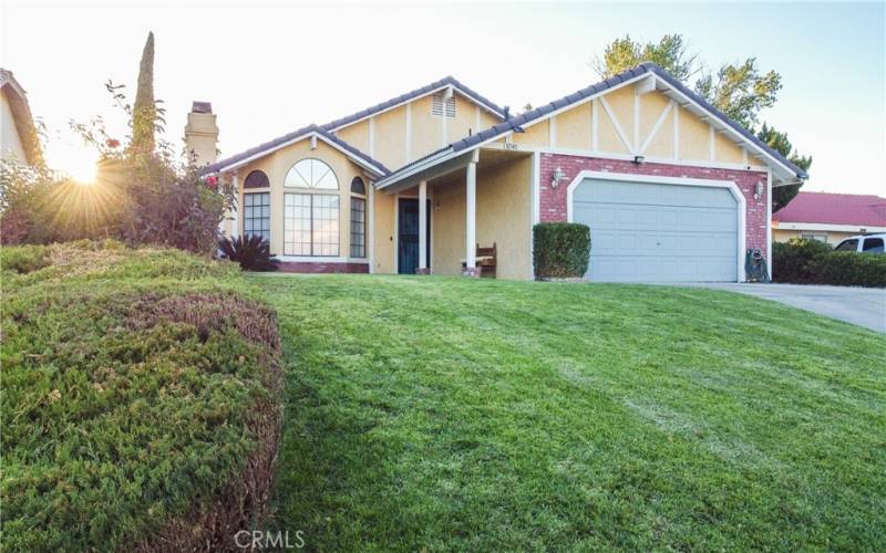 The front of the home showing grass and mature landscaping as well as brick walk way under the covered entry.  There is a tile roof and brick facade and the front door and garage are painted to match.