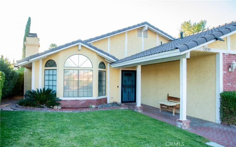 The front of the home showing grass and mature landscaping as well as brick walk way under the covered entry.  There is a tile roof and brick facade and the front door and garage are painted to match.