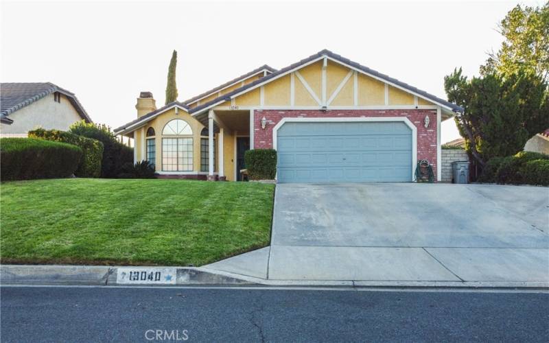 The front of the home showing grass and mature landscaping as well as brick walk way under the covered entry.  There is a tile roof and brick facade and the front door and garage are painted to match.