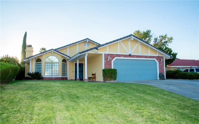 The front of the home showing grass and mature landscaping as well as brick walk way under the covered entry.  There is a tile roof and brick facade and the front door and garage are painted to match.