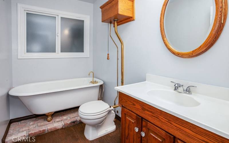 Guest bathroom done in a custom water closet design, rich wood-grained vanity, framed dressing mirror, vintage ball and claw freestanding tub, wood-grained vanity, dressing mirror, high tank pull chain commode, and wood floors.