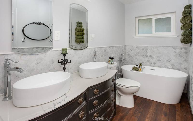 Remodeled main bathroom with custom-made antique quartz crowned vanity, dual vessel bowl basins, modern lighting, large free-standing soaking tub, and 4 ft. high marble tile wainscoting.