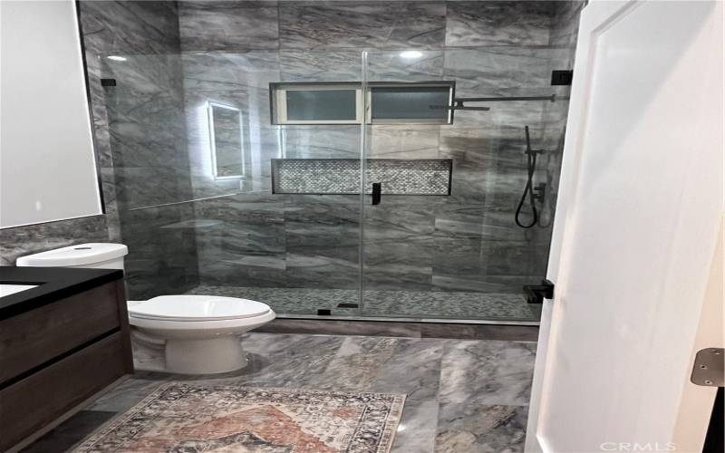 modern design with stunning dark marble tile throughout, creating a spa-like atmosphere. The spacious glass-enclosed shower is fitted with elegant brass fixtures and a stylish herringbone tile accent, blending functionality with high-end finishes.