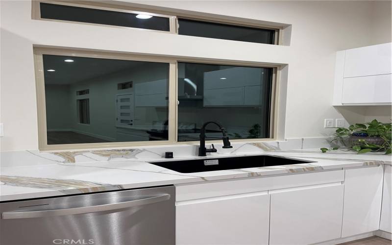 This close-up view of the kitchen in the newly built home highlights its modern, stylish design. The sleek marble-patterned countertops and backsplash add a touch of elegance, while the large black sink and high-end black faucet bring a bold contrast to the white cabinetry. A stainless steel dishwasher is seamlessly integrated, adding functionality without compromising the clean aesthetic. The large window above the sink invites natural light, enhancing the kitchen's bright and airy atmosphere.