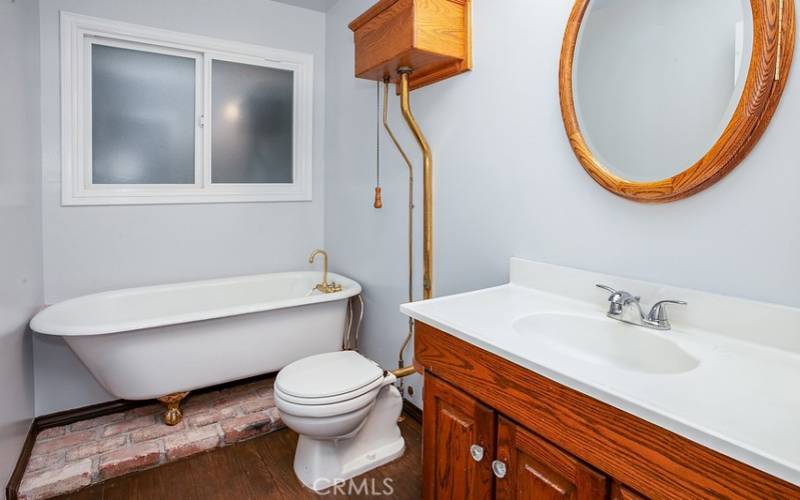 Guest bathroom done in a custom water closet design, rich wood-grained vanity, framed dressing mirror, vintage ball and claw freestanding tub, wood-grained vanity, dressing mirror, high tank pull chain commode, and wood floors.