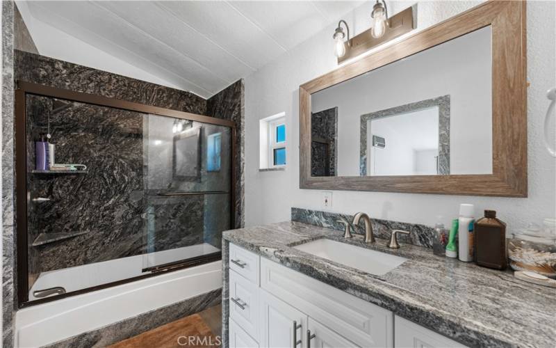 This bathroom features a modern design with a dark, stone-tiled shower and a double vanity. The granite countertop adds a luxurious touch, complemented by the rustic wooden-framed mirror and stylish light fixtures. It combines elegance with functionality, making it a standout feature.